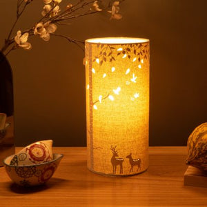 Image shows tube shaped beige fabric lamps with white Deers designs painted on and cut out shapes which a warm light shines out of.