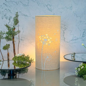 Image shows tube shaped beige fabric lamps with white Dandelion designs painted on and cut out shapes which a warm light shines out of.