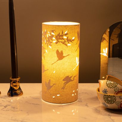 Image shows tube shaped beige fabric lamps with white Cranes and Bamboo designs painted on and cut out shapes which a warm light shines out of.