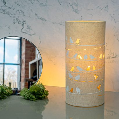Image shows tube shaped beige fabric lamps with white Birds on a wire designs painted on and cut out shapes which a warm light shines out of.