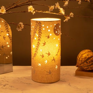 Image shows tube shaped beige fabric lamps with white Swallows at dawn designs painted on and cut out shapes which a warm light shines out of.